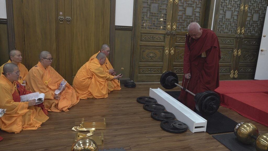 在報名考試的佛弟子們都上前單手提不起從一百八十磅起考的金剛杵失敗,已近九十的開初教尊竟然將之提起,甚至加碼到兩百磅亦依法懸提七秒過關,眾人驚嘆神力。圖中僧尼各司其職監考並紀錄考試結果。
